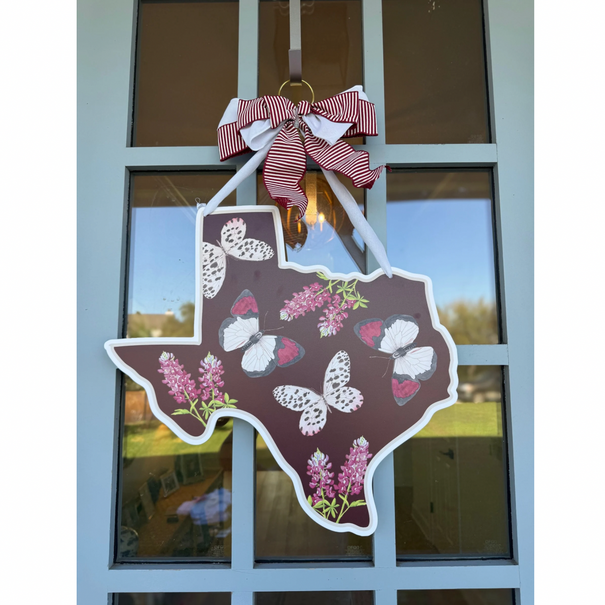 A decorative hanging sign shaped like the state of Texas, featuring white butterflies and pink flowers on a dark brown background, suspended from a red-and-white striped bow in front of a window.
